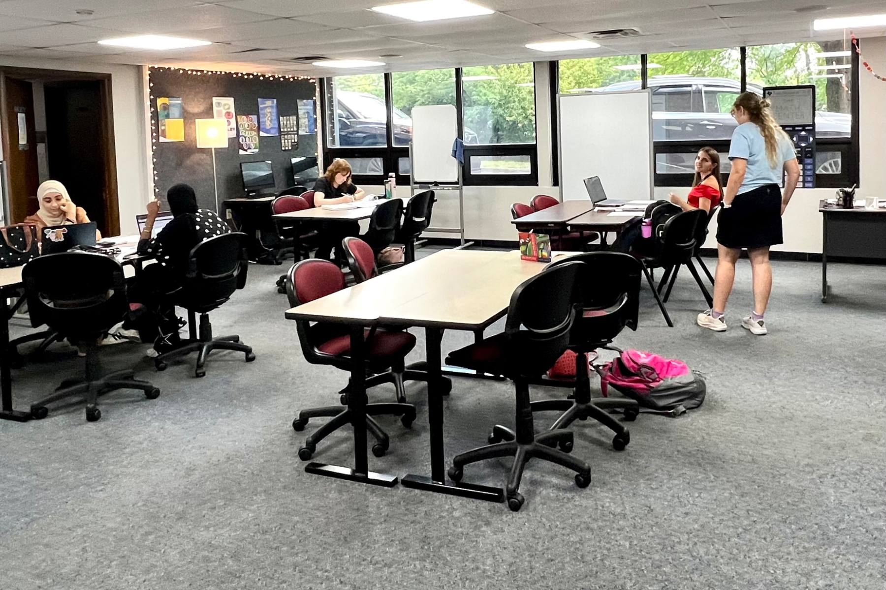 This image shows a bright room with windows in the background. There are four tables with student working independently and in pairs. There are computers, posters and whiteboards in the background.