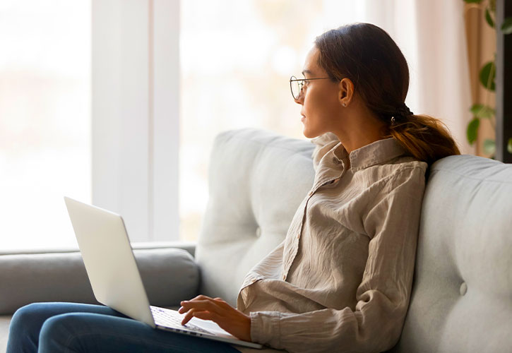 A young woman with a laptop looks contemplatively out a window
