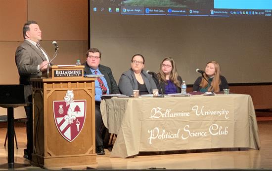 Speaker in front of a Bellarmine Podium next to four panelists