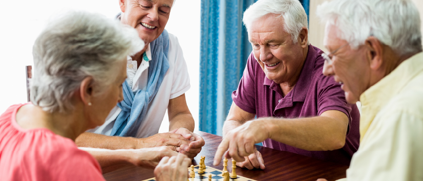 A group of senior citizens sit at a game table where two are playing chess